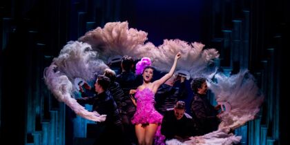 Scènefoto voor de musical Josephine B. met een zangeres in een roze jurk, omringt door dansers met verenwaaiers.